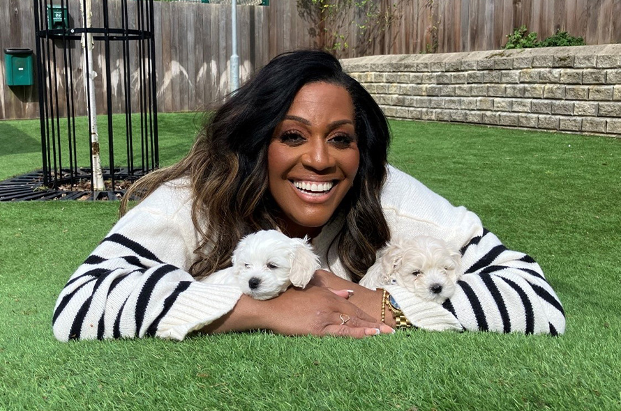 TV presenter laying on the grass with two small white puppies